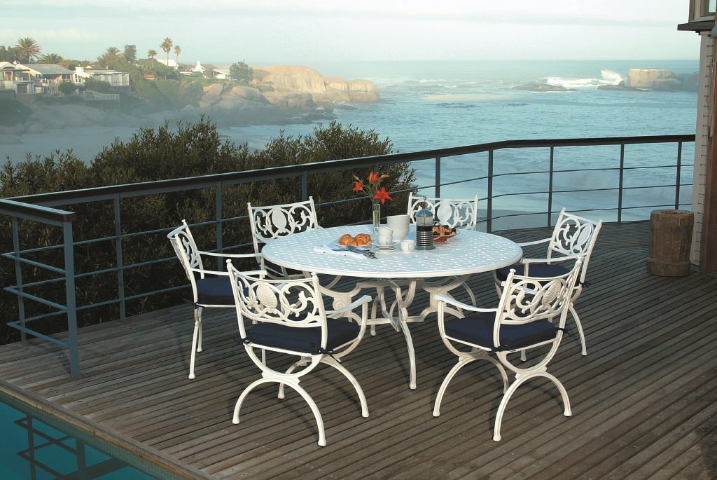 Artemis outdoor dining set by Oxley's Furniture, featuring elegant white cast aluminium chairs and a round table with navy cushions, displayed on a coastal deck with stunning ocean views—perfect for timeless luxury and outdoor living.